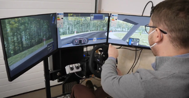 Zdjęcie przedstawia ucznia, który używa stymulatora do nauki jazdy. Urządzenie składa się z trzech ekranów i kierownicy.