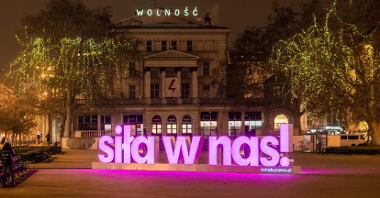 Zdjęcie przedstawia świecący na różowo przestrzenny napis "siła w nas". W tle budynek Arkadii.