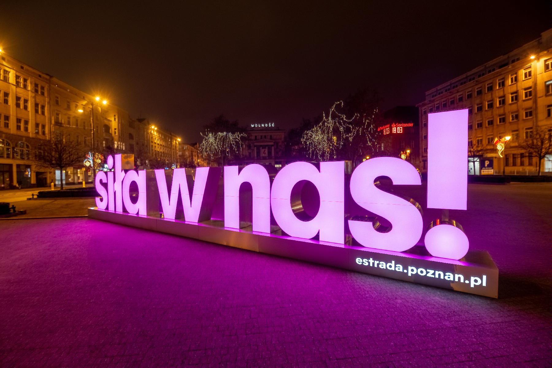 Zdjęcie przedstawia świecący na różowo przestrzenny napis "siła w nas". W tle plac Wolności i budynek Arkadii. - grafika artykułu
