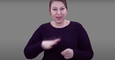 Kadr z filmu: Eunika Lech, tłumaczka polskiego języka migowego, miga, stojąc na szarym tle