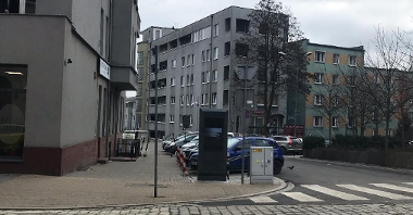 Zdjęcie przedstawia multimedialny totem, stojący na chodniku, w sąsiedztwie jezdni i zaparkowanych samochodów. W tle widać bloki.