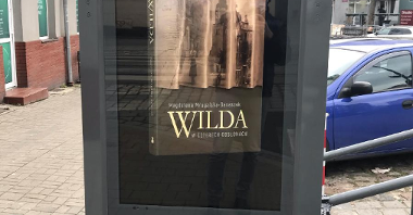 Zdjęcie przedstawia multimedialny totem. Na ekranie wyświetlona jest okładka książki pt."Wilda w czterech odsłonach". W tle widać zaparkowane samochody.