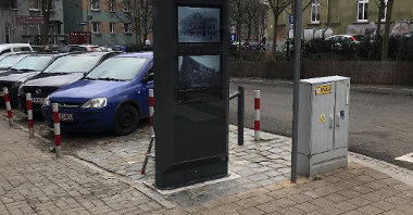 Zdjęcie przedstawia multimedialny totem. Urządzenie stoi na chodniku. Obok znajdują się zaparkowane na poboczu jezdni samochody.