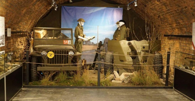 Zdjęcie przedstawia muzealne eksponaty: samochód bojowy, działko strzelnicze oraz manekiny ubrane w żołnierskie stroje.