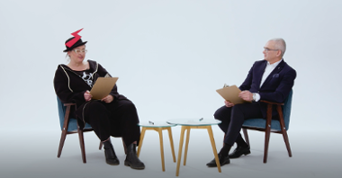 Kard z filmu: na krzesłach, przy stoliku siedzą kobieta i mężczyzna; kobieta ma symbol Strajku Kobiet na kapeluszu, mężczyzna jest ubrany w garnitur