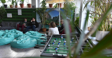 Zdjęcie przedstawia strefę relaksu w szkole. Widać na nim uczniów siedzących na kanapach, a także pufy, stół do gry w piłkarzyki, donice z roślinami oraz plakat przedstawiający jezioro i góry.