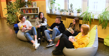 Strefa relaksu w szkole. Zdjęcie przedstawia uczniów siedzących na pufach na szkolnym korytarzu. Za ich plecami widać biblioteczkę z książkami oraz doniczki z kwiatami, stojące na parapecie.