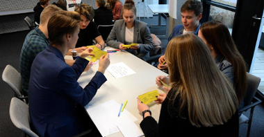 Zdjęcie przedstawia uczniów siedzących przy stołach. Młodzież trzyma w ręku żółte karteczki, na stole leża kolorowe pisaki.