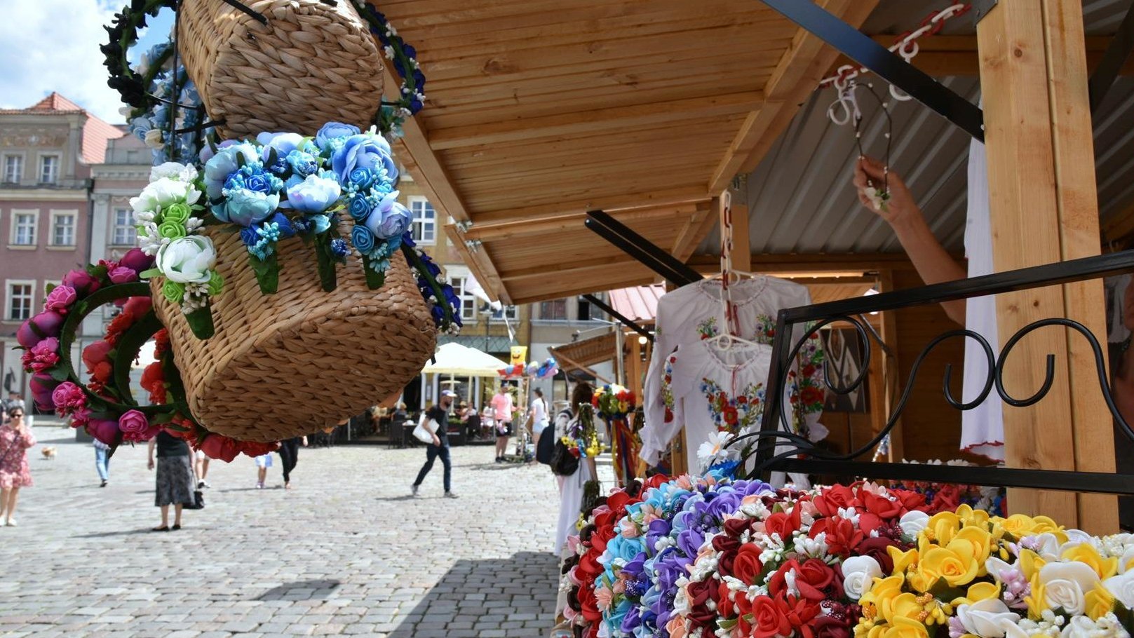 Zdjęcie przedstawia drewniany domek handlowy stojący na Starym Rynku. Na stoisku znajdują się bukiety ze sztucznych kwiatów (niektóre umieszczone zostały w wiklinowych koszykach) oraz bluzki w kwiatki. - grafika artykułu
