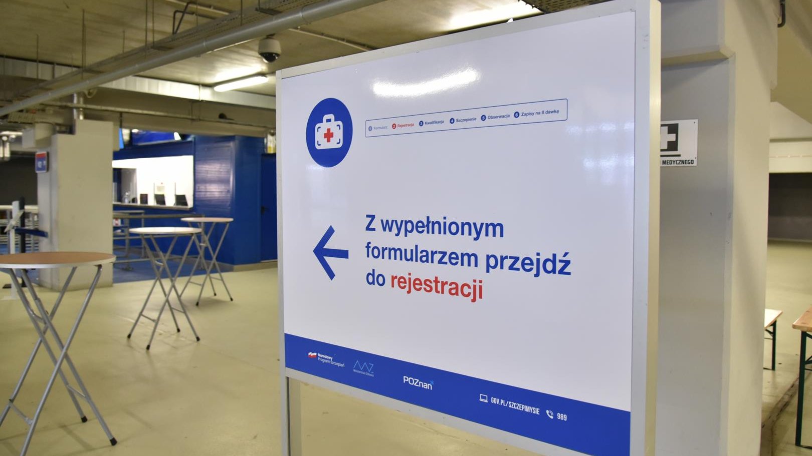 Wnętrze punktu, na pierwszym planie baner wskazujący drogę do rejestracji, w tle stoliki - grafika artykułu