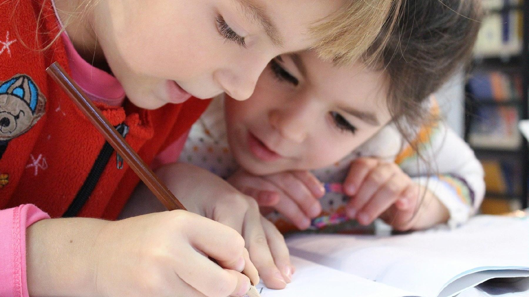 Zdjęcie przedstawia dwójkę dzieci pochylonych nad książką. Jedno z dzieci trzyma w ręku ołówek i coś pisze. - grafika artykułu