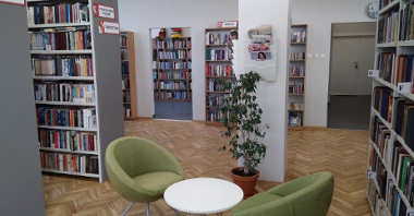 Zdjęcie przedstawia wnętrze biblioteki. Widać na nim półki z książkami oraz mały biały stoliczek z dwoma zielonymi krzesłami.