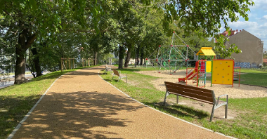 Na zdjęciu widać alejkę spacerową biegnącą wśród zieleni. Obok znajduje się plac zabaw dla dzieci oraz ławki.