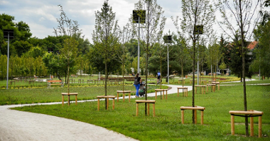 Zdjęcie przedstawia teren zielony - trawę, drzewa, krzewy. Przez park ciągną się alejki spacerowe, po których chodzą ludzie, np. matka z dzieckiem w wózku. Jeżdżą po nich także rowerzyści. Obok stoją ławki.