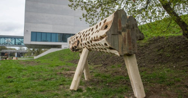 Zdjęcie przedstawia drewnianą instalację w kształcie pnia z otworami dla ptaków i owadów. W tle widać budynek Bramy Poznania.