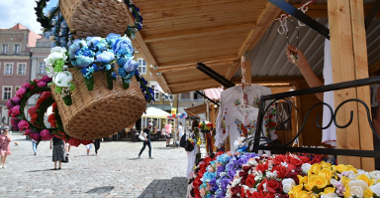 Zdjęcie przedstawia drewniany domek handlowy, stojący na Starym Rynku. Na stoisku widać kwiaty w koszu wiklinowym oraz białe bluzki w kwiaty. W tle można dostrzec ludzi.