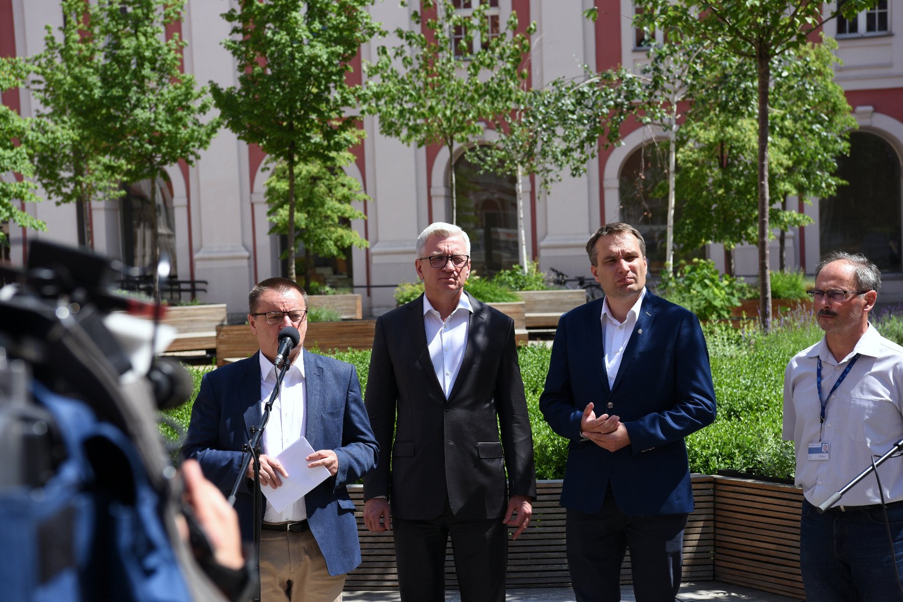 Na zdjęciu dziedziniec urzędu miasta, przy mikrofonach stoi czterech mężczyzn, w tym Jacek Jaśkowiak i Mariusz Wiśniewski, w tle drzewa - grafika artykułu