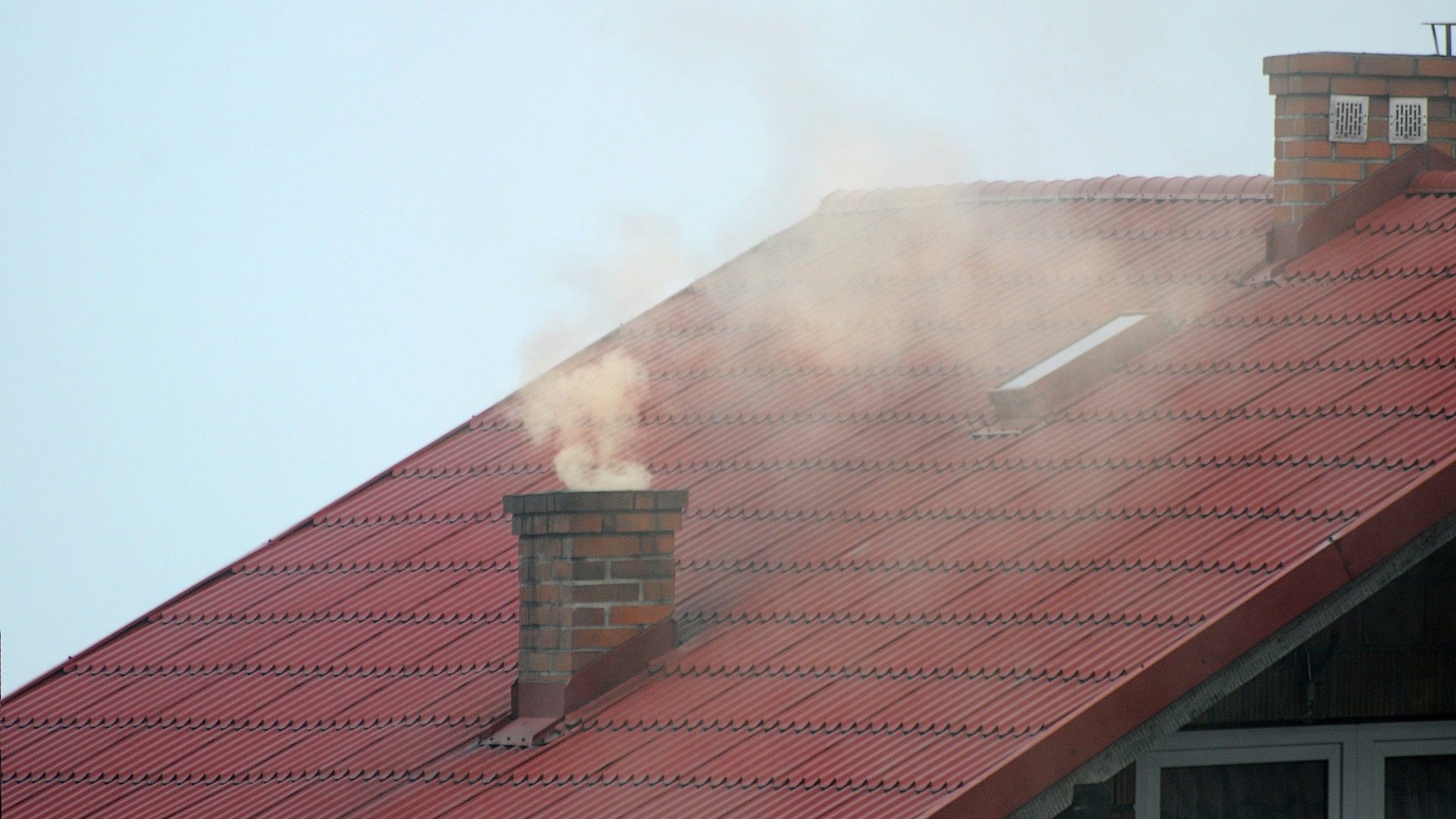 Na zdjęciu zbliżenie na czerwony dach, w centrum dymiący komin - grafika artykułu