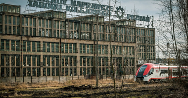 Na zdjęciu stary, zrujnowany budynek po ZNTK, przed nim przejeżdża nowoczesny pociąg