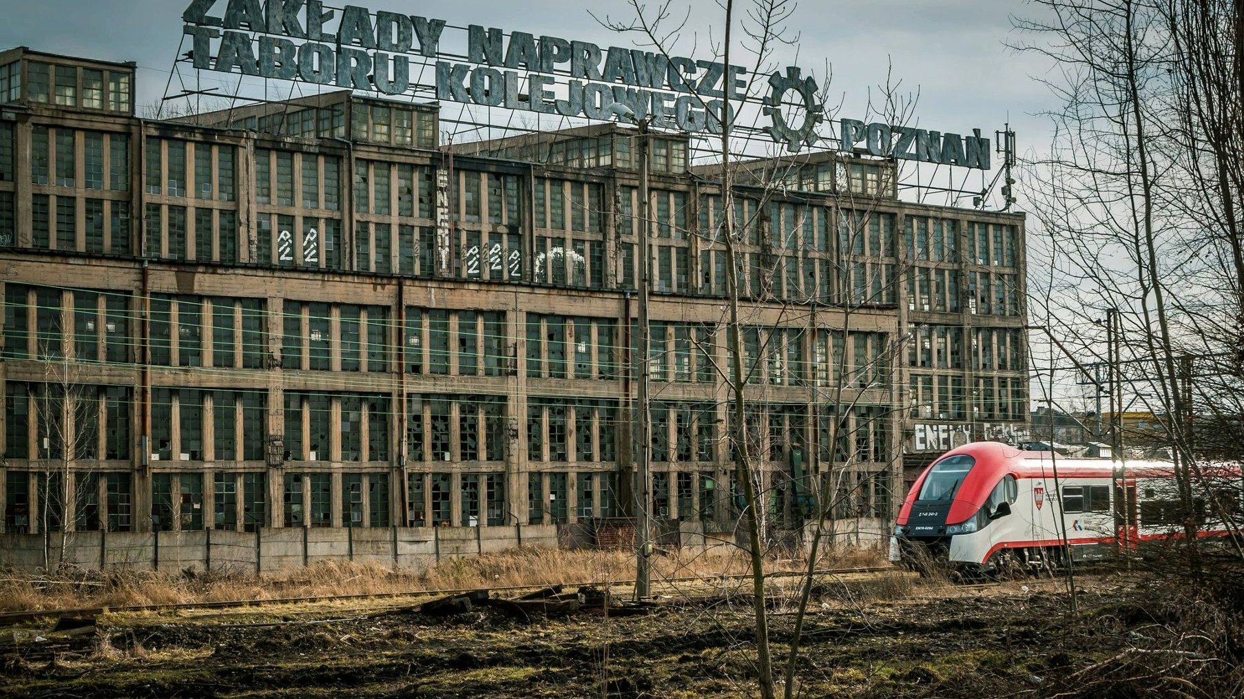 Na zdjęciu stary, zrujnowany budynek po ZNTK, przed nim przejeżdża nowoczesny pociąg - grafika artykułu