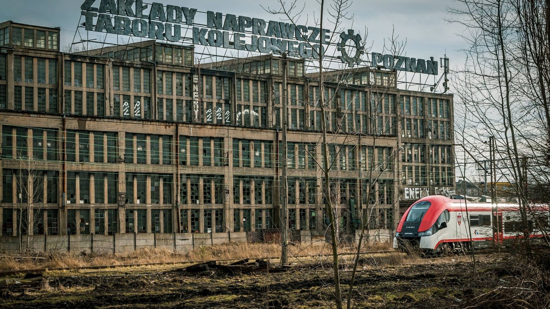 Na zdjęciu stary, zrujnowany budynek po ZNTK, przed nim przejeżdża nowoczesny pociąg