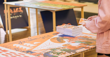 Zdjęcie przedstawia kolorową ilustrację leżącą na stoliku, obok znajdują się kolorowe pocztówki. Przy stoliku stoi osoba w różowej kurtce, trzymająca jedną z pocztówek w ręce. W tle widać drugi stolik.