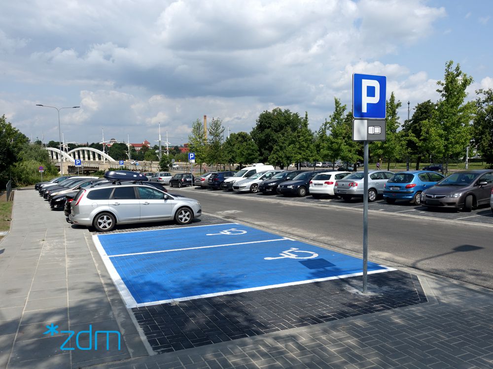 Na pierwszym planie parking przy ulicy, miejsce dla osób z niepełnosprawnościami, w tle most św. Rocha - grafika artykułu