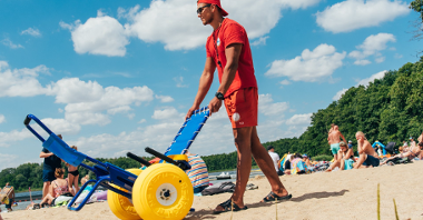 Zdjęcie przedstawia ratownika z wózkiem na plaży. Pcha on przed sobą wózek amfibie.