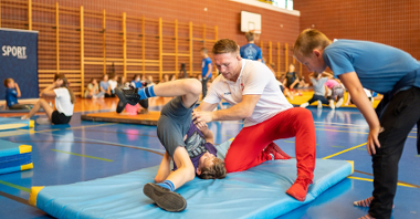 Na zdjęciu sala gimnastyczna i medalista olimpijski trenujący ucznia na macie, obok inne dzieci