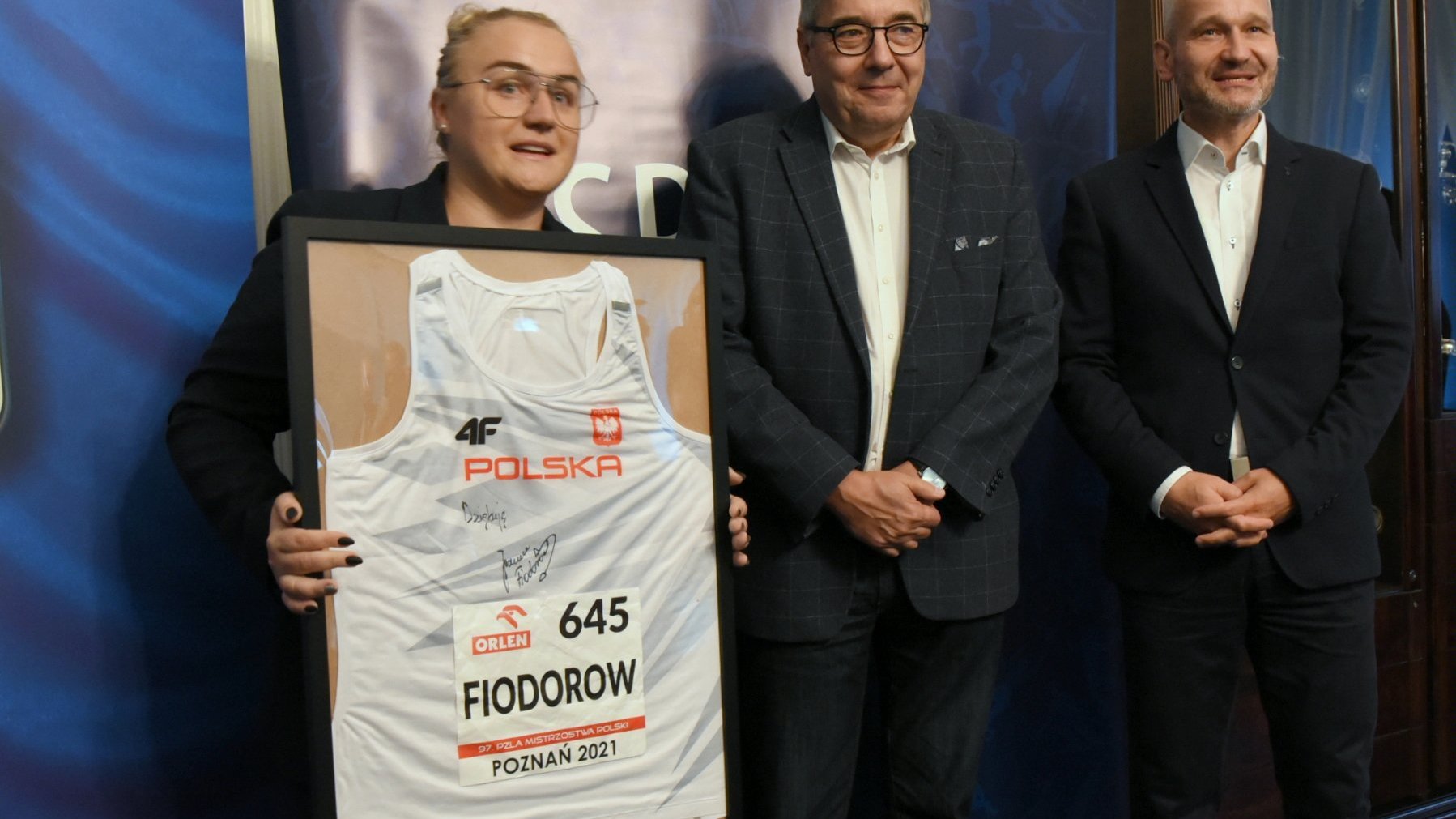 Na zdjęciu Joanna Fiodorow trzymająca oprawioną w ramkę swoją koszulkę, obok Grzegorz Ganowicz i Jędrzej Solarski