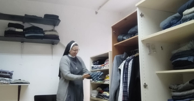 Na zdjęciu uśmiechnięta zakonnica składa męską koszulę, by ułożyć ją na regale pełnym ubrań