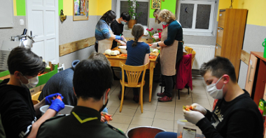 Na zdjęciu grupa wolontariuszy w niewielkim pomieszczeniu - kroją warzywa i przygotowują posiłki