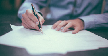 Zdjęcie przedstawia ręce mężczyzny trzymającego długopis i piszący coś na kartce papieru.