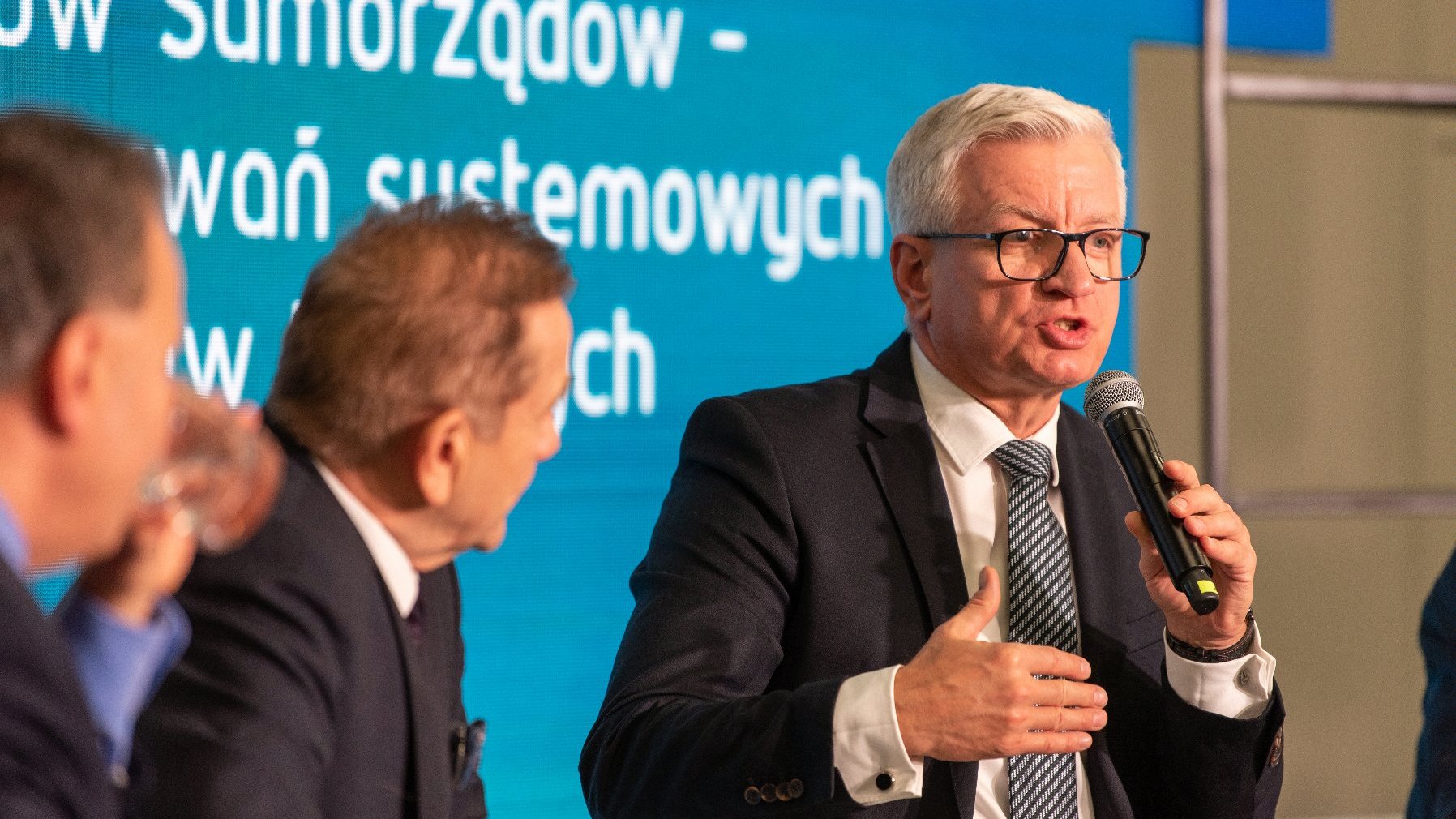 Jacek Jaśkowiak, prezydent Poznania, z mikrofonem w ręku, podczas debaty, w tle ekran z jej tytułem - grafika artykułu