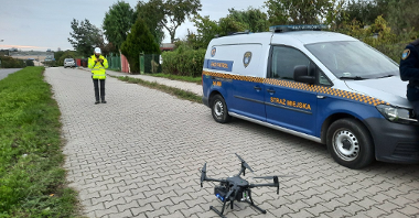 Zdjęcie przedstawia stojącego na chodniku drona, osobę, która go obsługuje oraz samochód straży miejskiej.