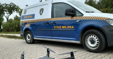 Zdjęcie przedstawia stojącego na chodniku drona oraz samochód straży miejskiej.