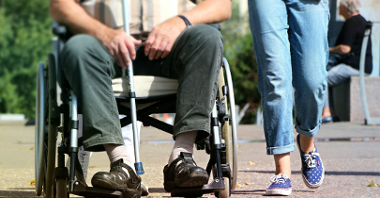 Zdjęcie przedstawia dwie osoby, jedna z nich znajduje się na wózku inwalidzkim. Na fotografii jest zbliżenie na nogi.
