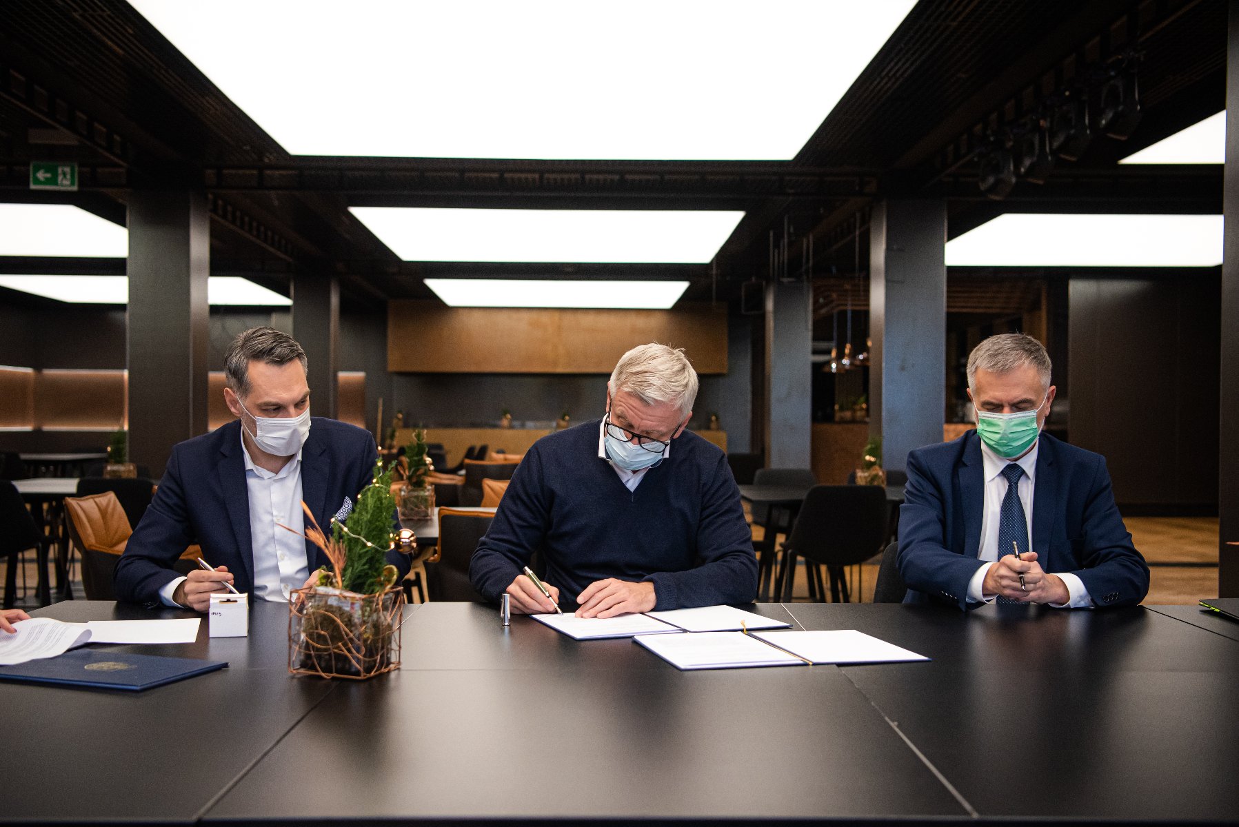 Zdjęcie przedstawia trzech mężczyzn za stołem. W środku znajduje się prezydent Poznania podpisujący umowę. - grafika artykułu