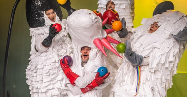 Zdjęcie przedstawia aktorów przebranych w białe stroje, wyglądające jak wykonane z papieru. W rękach trzymają kolorowe piłki.