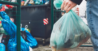 Zdjęcie przedstawia osobę niosącą worek ze śmieciami. Widać na nim jednak tylko rękę z workiem. W tle znajduje się kontener ze śmieciami.