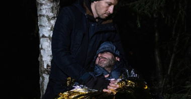 Zdjęcie przedstawia mężczyznę trzymającego drugiego mężczyznę. Ten drugi jest nieprzytomny, owinięty w folię termiczną.