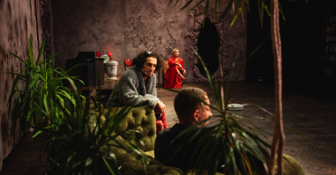 Zdjęcie przedstawia aktorów na scenie. Widać na nim siedzących dwóch mężczyzn i jedną kobietę.
