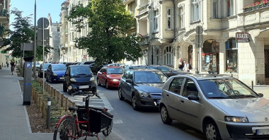 Na zdjęciu poznańska ulica, na niej samochody, na pierwszym planie chodnik i zaparkowany na nim rower