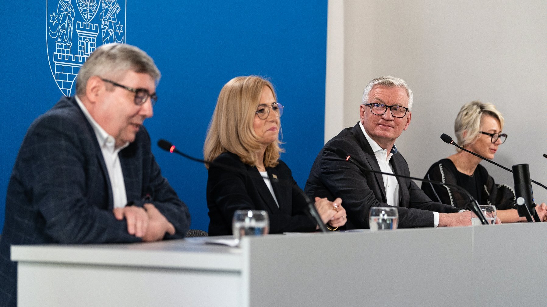 Na zdjęciu cztery osoby za stołem konferencyjnym, w środku prezydent Poznania, w tle granatowa ścianka - grafika artykułu