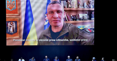 Zdjęcie przedstawia uczestników obrad siedzących przy stole oraz telebim, na którym widać Witalija Kliczko, mera Kijowa.