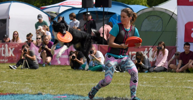 Zdjęcie przedstawia dziewczynę rzucającą frisbee oraz psa, który łapie frisbee w locie. W tle widać obserwujących pokaz ludzi.