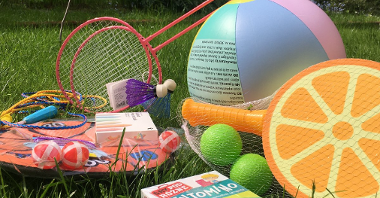 Na zdjęciu zestaw leżących na trawie zabawek, wśród nich piłka, paletki, lotki i gra planszowa