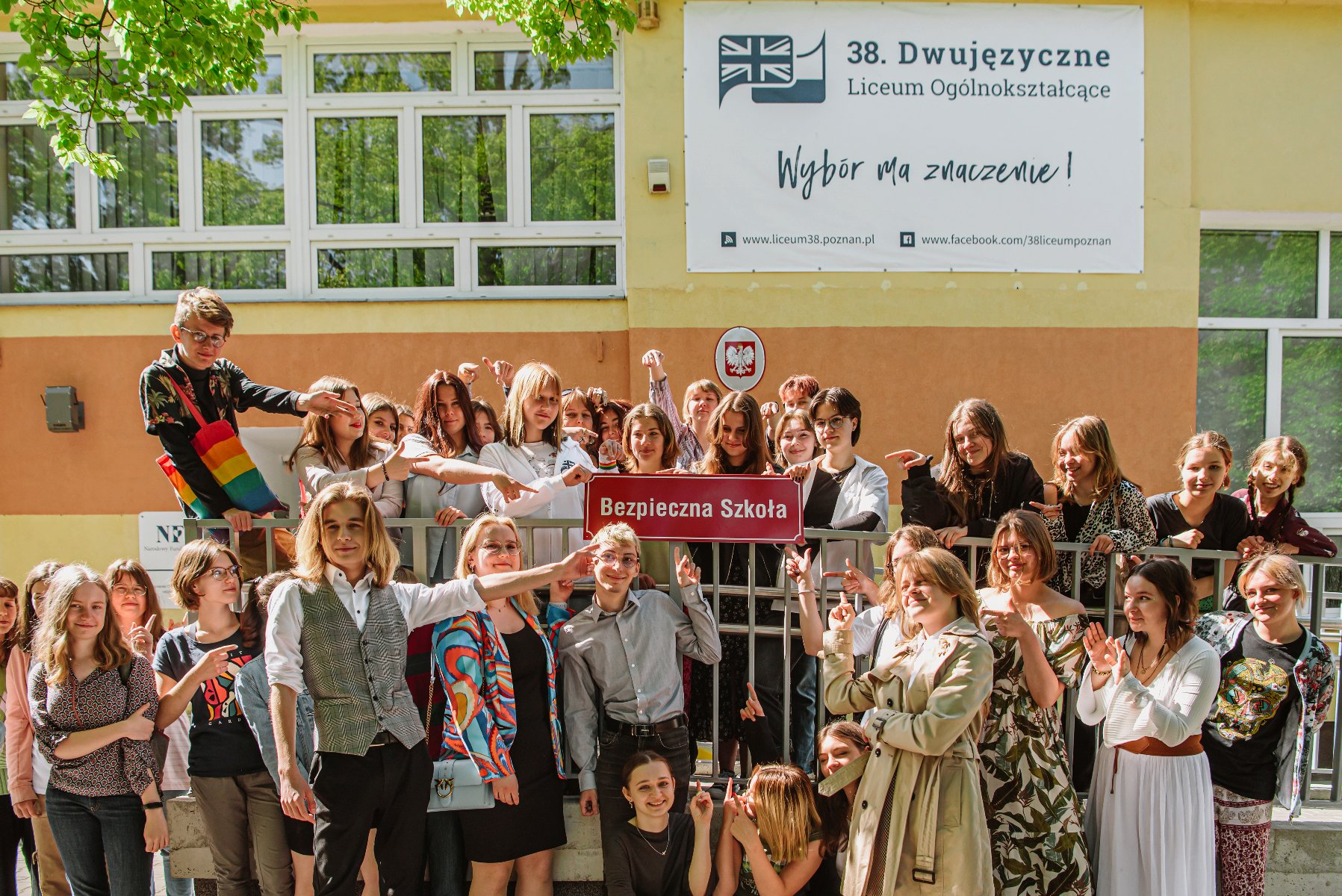 Grupowe zdjęcie uczniów i nauczycieli przed szkołą, pozujących z tabliczką z napisem "Bezpieczna szkoła". - grafika artykułu