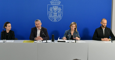 Na zdjęciu cztery osoby: dwóch mężczyzn i dwie kobiety siedzą za stołem konferencyjnym, za nimi granatowa ścianka z herbem miasta