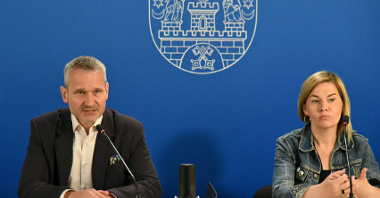 Na zdjęciu mężczyzna i kobieta za stołem konferencyjnym, za nimi gratatowa ścianka z herbem miasta